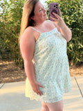A woman taking a mirror selfie outdoors wearing a light green and white floral print spaghetti-strap dress.