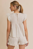 A woman shown from the back wearing a cream-colored plaid romper with ruffled sleeves and shorts, set against a solid beige background.
