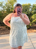 A woman takes a mirror selfie outdoors wearing a light blue and white floral print spaghetti-strap dress. The background shows green foliage and a concrete walkway.