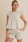 A woman modeling a matching two-piece set featuring a ruffled sleeveless top and shorts in a beige and white gingham plaid pattern, set against a solid beige background.